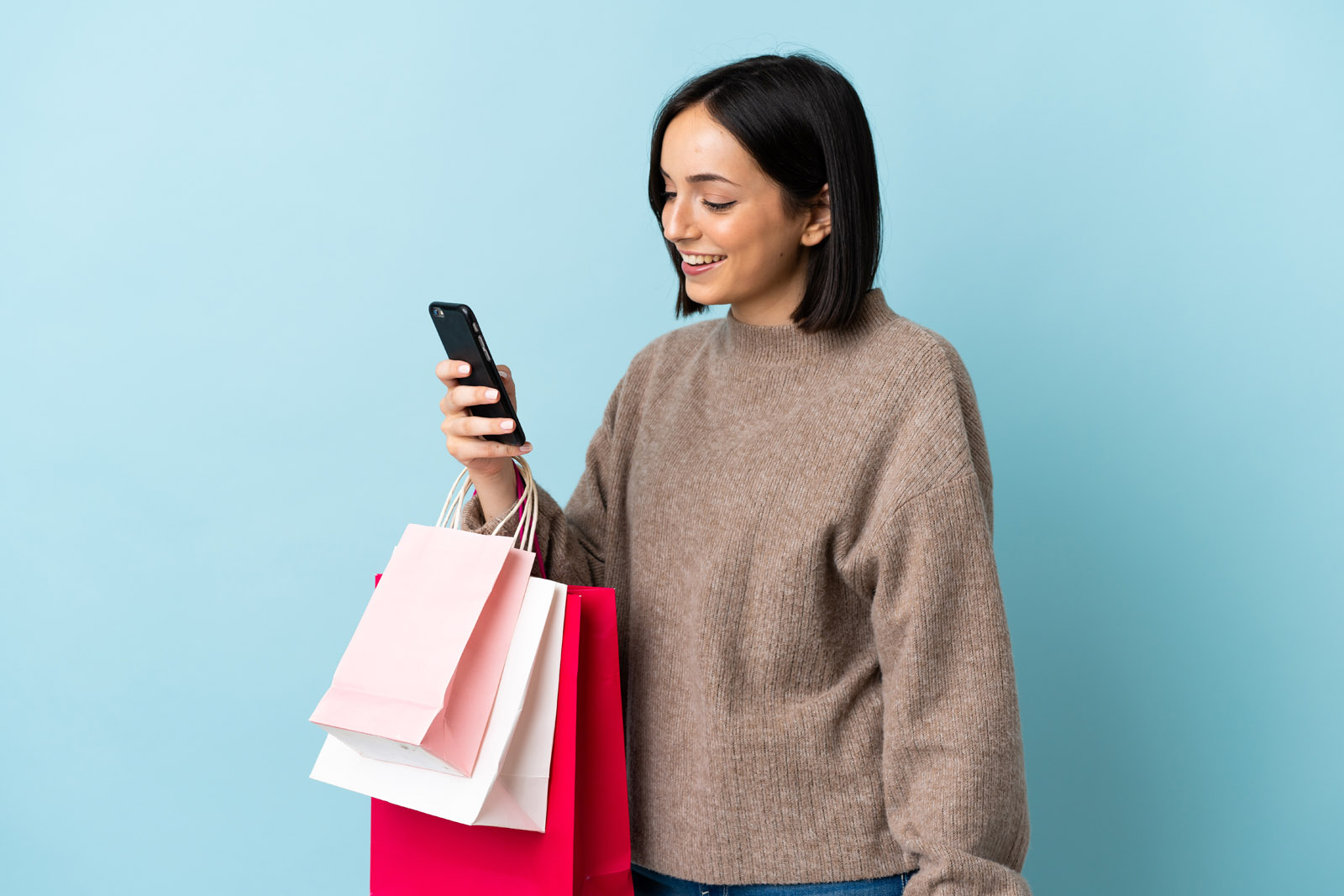 Young caucasian woman isolated on blue background holding shopping bags and writing a message with her cell phone to a friend